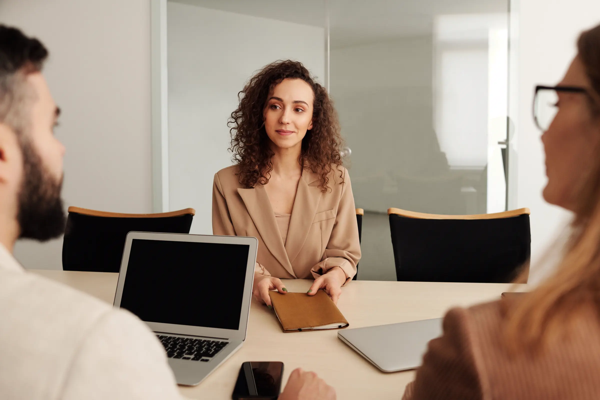 A candidate in a camel blazer at an interview, across the table from two interviewers in a bright office.