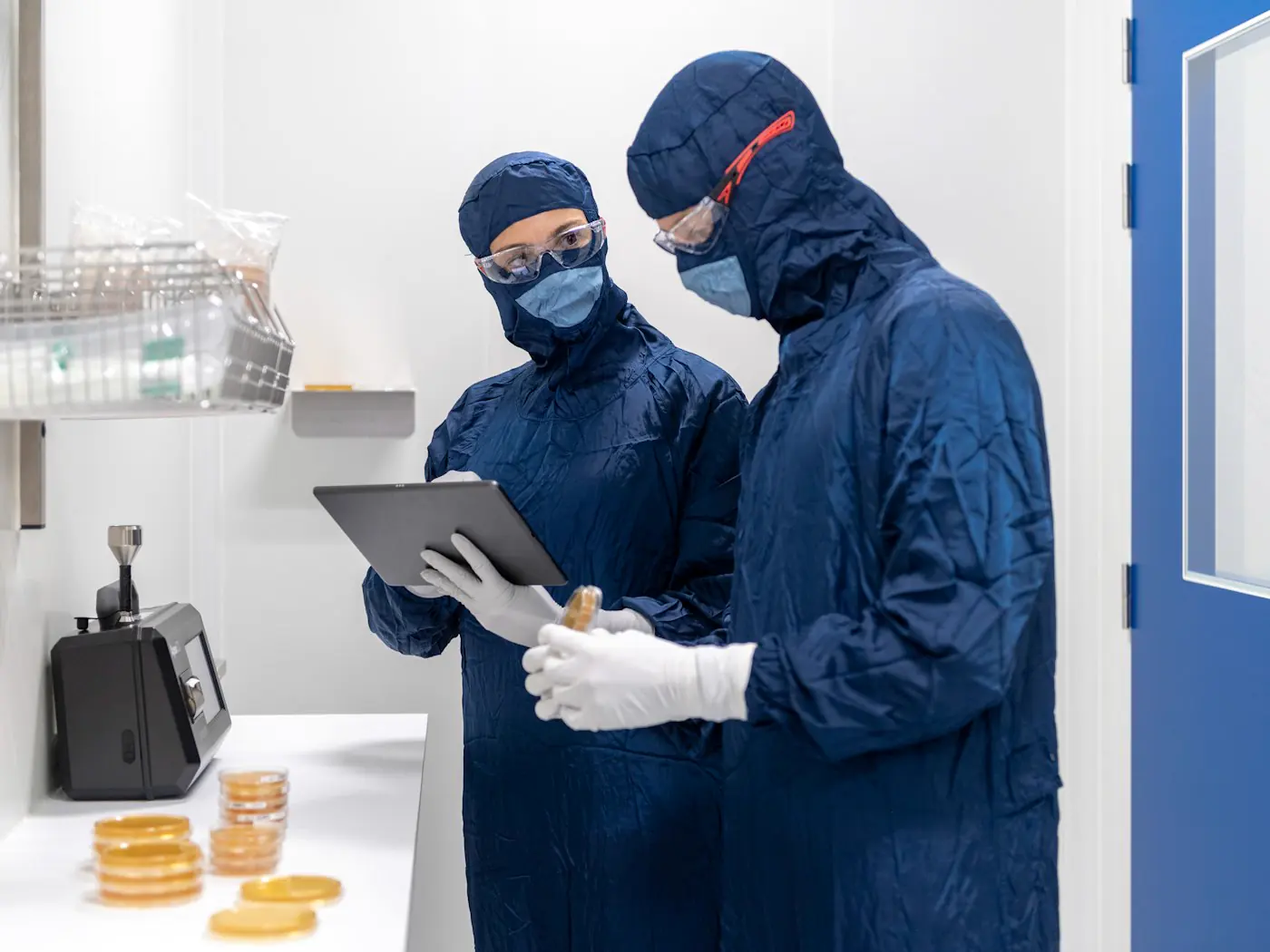 Two operators in navy cleanroom gowns and goggles checking a tablet on a production floor.