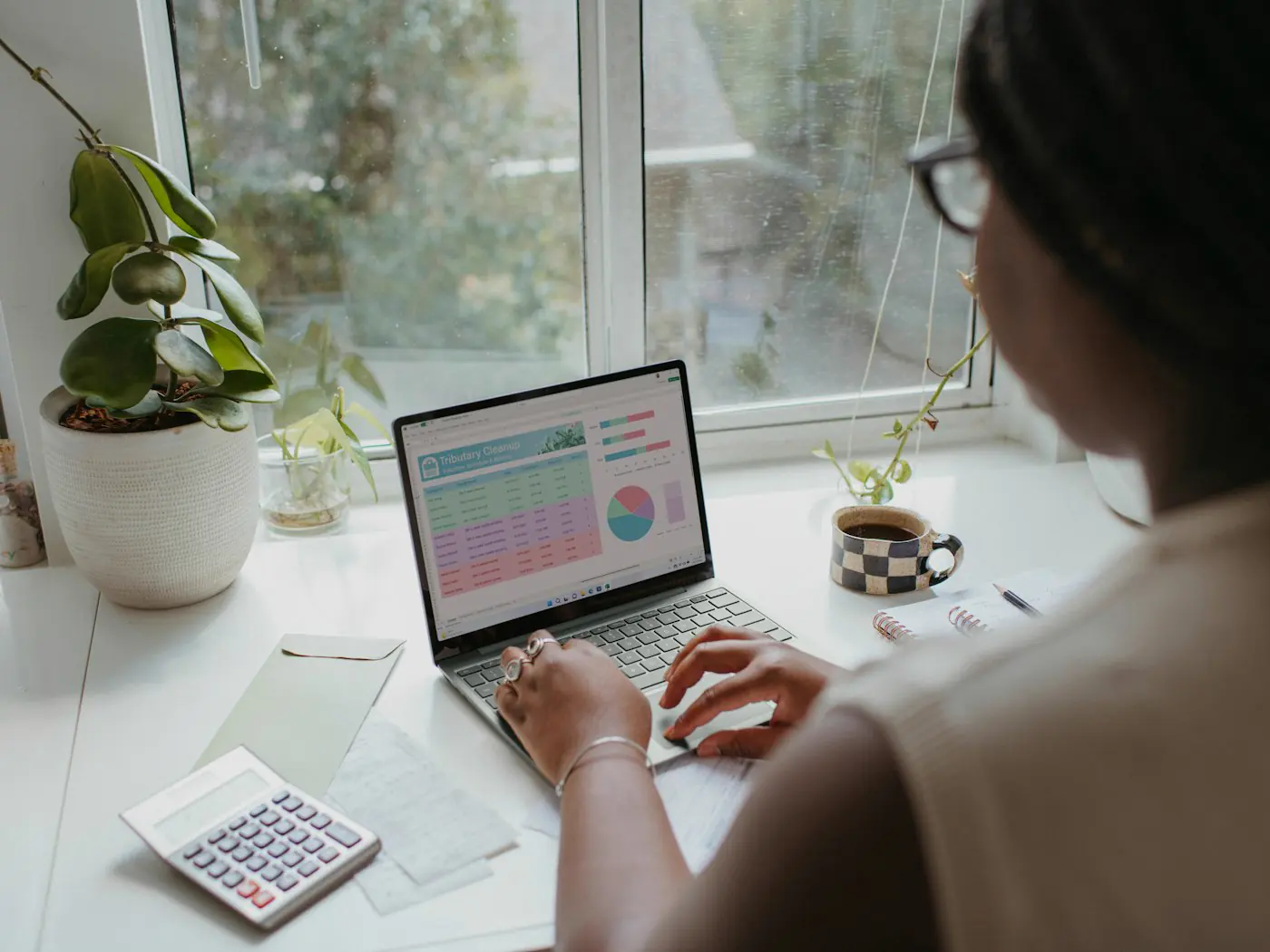 An analyst reviewing a spreadsheet and charts on a laptop at a desk by a window.